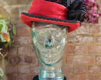 May include: A black and red top hat with a red band and black feather accent. The hat has a velvet texture and is displayed on a glass head form. The hat is trimmed with black decorative trim.