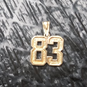 May include: Gold-colored pendant featuring the number "83" in a block letter style. The pendant has a loop at the top for a chain. The background is a textured, dark surface.