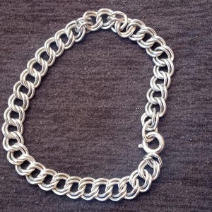 May include: A silver-toned chain bracelet constructed from interlocking double circular links. The bracelet features a circular spring ring clasp mechanism that connects the two ends of the chain, creating a continuous loop of metallic links.