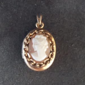 May include: An oval gold-toned metal locket pendant featuring a central cameo with a white profile silhouette of a person against a light brown background. The locket is surrounded by a decorative twisted rope-style border and has a small bail at the top.