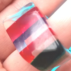 May include: A colorful, rectangular ring with a clear base. The ring features horizontal stripes of red, purple, pink, and green. The ring is held by a person.