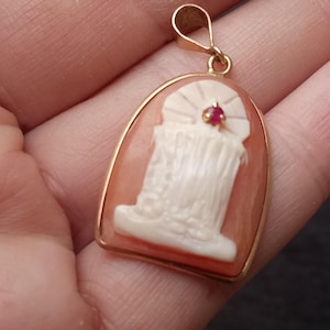 May include: A gold pendant featuring a carved white shell cameo set against a peach-colored background. The cameo depicts a vertical figure with a small, round, red gemstone accent at the top. The pendant has a gold bail at the top for a chain.