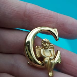 May include: A gold-colored brooch featuring an angel seated on a crescent moon. The angel has detailed wings and is in a seated position. The brooch has a polished finish and is held in a hand against a turquoise background.