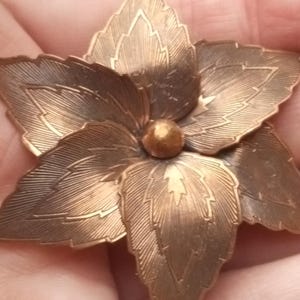 May include: A detailed, bronze-colored flower brooch with six detailed petals and a central sphere. The brooch is held in a hand, showcasing its intricate design and vintage aesthetic. The leaves have detailed veins.