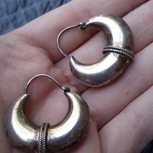May include: A pair of silver-toned hoop earrings featuring a thick, crescent-shaped body with a smooth, polished finish and a central decorative band of twisted rope-like texture. Each earring includes a thin, curved wire closure mechanism.