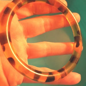 May include: A translucent, round bangle bracelet with a tortoiseshell pattern. The bracelet features alternating bands of amber and dark brown, with a glossy finish. The bangle is held against a light orange background.