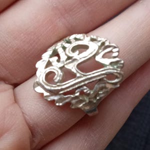 May include: Silver-toned filigree ring with an openwork design. The ring features an ornate, swirling pattern with floral elements. The ring is displayed on a finger, showcasing its intricate details and craftsmanship.