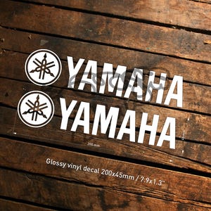 Op de afbeelding: Twee witte Yamaha-stickers op een houten oppervlak. Elke sticker bevat het Yamaha-logo en het woord "YAMAHA" in een vetgedrukte, schreefloze letter. De stickers zijn 200 mm lang en 45 mm hoog. De tekst "Glossy vinyl decal 200x45mm" is ook zichtbaar.