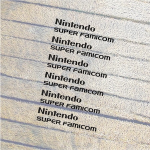 Könnte beinhalten: Nahaufnahme einer weißen Oberfläche mit dem Text "Nintendo SUPER FAMICOM", der sechsmal wiederholt wird.