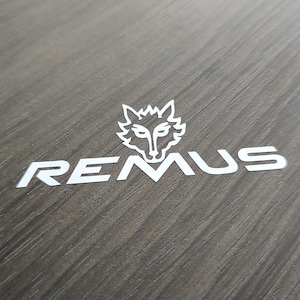 以下が含まれることがあります： 暗い木製の表面に白いロゴ。ロゴは、スタイリッシュなオオカミの頭と白い文字で「REMUS」という単語を特徴としています。