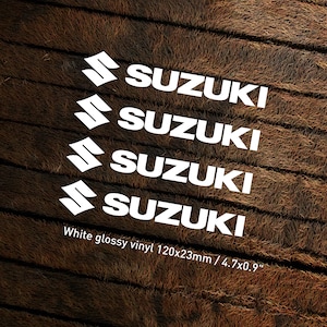 May include: Four white glossy vinyl decals with the word "SUZUKI" in a bold, sans-serif font. The decals are arranged in a staggered pattern, with each decal slightly offset from the one above it. The decals are on a brown, textured background. White glossy vinyl 120x23mm / 4.7x0.9"