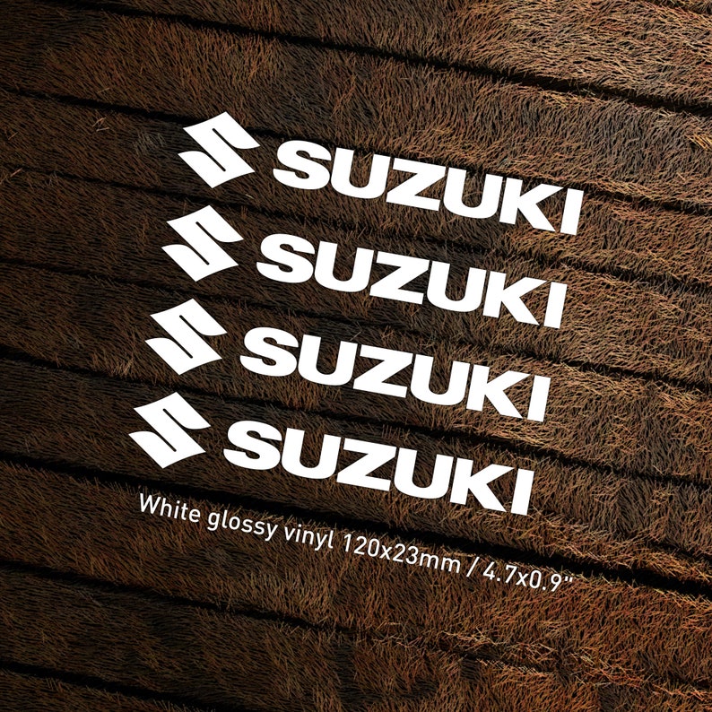 May include: Four white glossy vinyl decals with the word "SUZUKI" in a bold, sans-serif font. The decals are arranged in a staggered pattern, with each decal slightly offset from the one above it. The decals are on a brown, textured background. White glossy vinyl 120x23mm / 4.7x0.9"