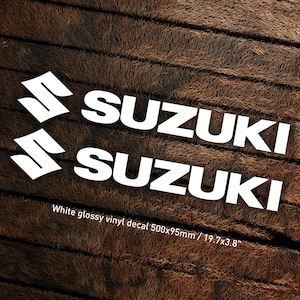 May include: Two white vinyl decals with the word "SUZUKI" in bold white letters. The decals are on a brown woodgrain background. White glossy vinyl decal 500x95mm / 19.7x3.8"