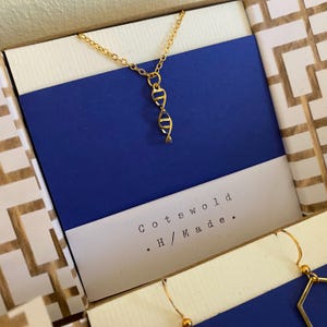 May include: A gold-coloured necklace with a DNA helix pendant is presented in an open gift box. The box has a blue background and a white card with the text "Cotswold H/Made." The box is decorated with a geometric pattern.
