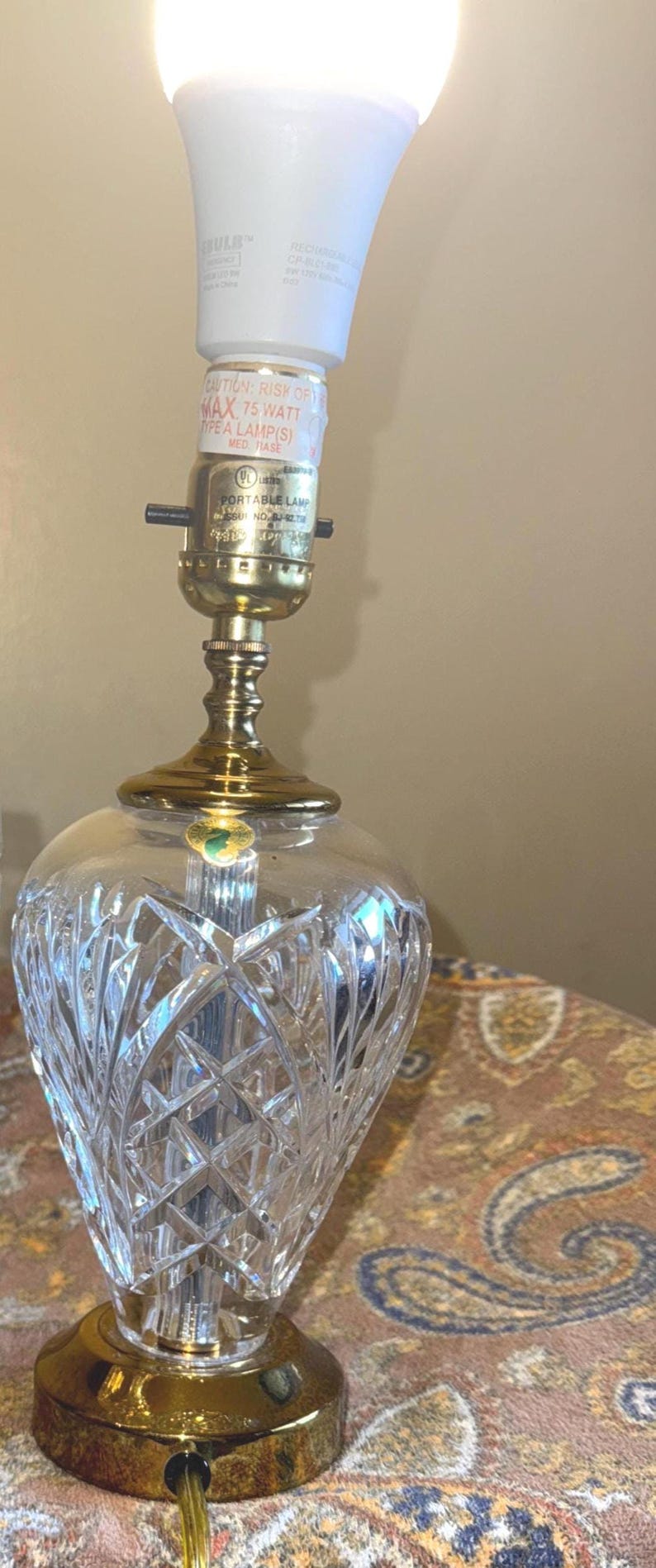May include: A crystal table lamp with a cut glass shade and a brass base. The lamp has a white LED bulb and a label that reads "CAUTION: RISK OF FIRE MAX 75 WATT TYPE A LAMP(S)".