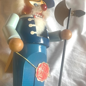 May include: A wooden nutcracker figure dressed in a blue uniform with a matching top hat featuring a yellow flower emblem. The figure has a white beard, holds a metal axe, and wears a red tag with the text Steinbach Volkskunst.