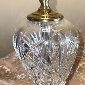 May include: A clear crystal table lamp with a gold-colored base and accents. The lamp features a cut glass design on the bulbous body and a brass-colored top. The lamp is designed for indoor use.