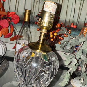 May include: A crystal table lamp with a gold-colored base and accents. The lamp has a clear, faceted glass shade and a cylindrical lamp holder. The lamp is next to a wine bottle and decorative foliage.