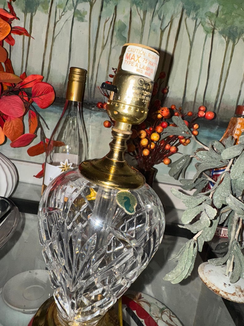 May include: A crystal table lamp with a gold-colored base and accents. The lamp has a clear, faceted glass shade and a cylindrical lamp holder. The lamp is next to a wine bottle and decorative foliage.