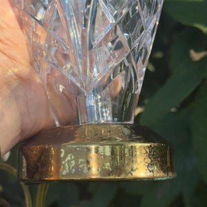 May include: A clear crystal vase with a geometric pattern. The vase sits on a gold-colored metal base. The base has a slightly tarnished appearance. The vase is held in front of a green background.