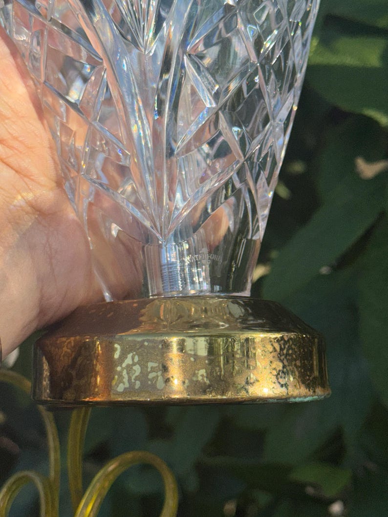 May include: A clear crystal vase with a geometric pattern. The vase sits on a gold-colored metal base. The base has a slightly tarnished appearance. The vase is held in front of a green background.