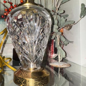 May include: A clear crystal table lamp with a gold-colored base and top. The lamp has a cut glass design and a brass-colored light fixture. The lamp is on a glass shelf with decorative items in the background.