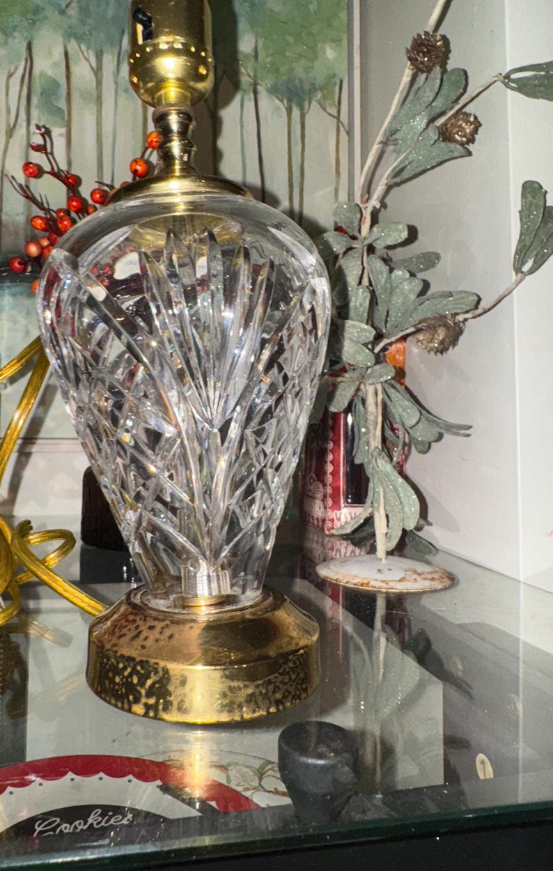 May include: A clear crystal table lamp with a gold-colored base and top. The lamp has a cut glass design and a brass-colored light fixture. The lamp is on a glass shelf with decorative items in the background.