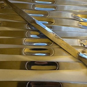 May include: A set of ten silver-toned metal butter knives with rounded handles. The knives are arranged in a stack, with the handles facing up. The brand name "Christofle" is visible on one of the knives.