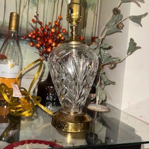 May include: A crystal table lamp with a brass base and top. The lamp has a cut glass design and a gold-colored electrical cord. The lamp is on a glass surface.