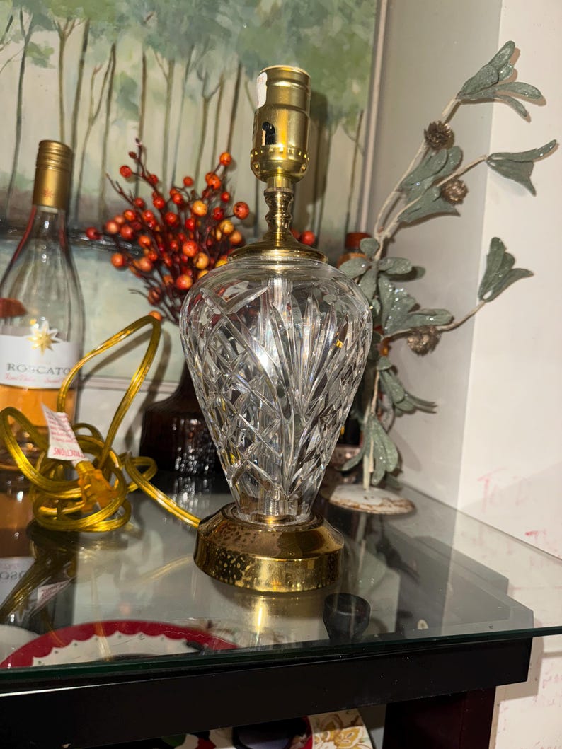 May include: A crystal table lamp with a brass base and top. The lamp has a cut glass design and a gold-colored electrical cord. The lamp is on a glass surface.