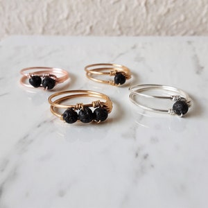 May include: Four wire-wrapped rings with black lava beads. The rings are in rose gold, gold, and silver tones. The rings are displayed on a white and grey marble surface.