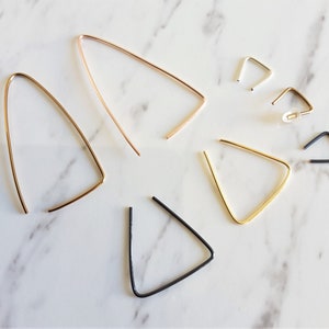 May include: A collection of minimalist earrings in gold, rose gold, silver, and black finishes. The earrings display geometric shapes, including triangles and elongated teardrops, with a modern and elegant design. The jewellery is presented on a white marble surface.