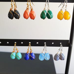May include: Eight pairs of teardrop-shaped earrings displayed on a black stand. The earrings feature smooth, solid-coloured stones wrapped in thin metal wire, including black, coral, green, yellow, turquoise, blue, light blue, and grey colours.