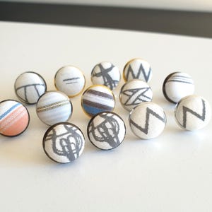 May include: A collection of thirteen circular buttons featuring fabric-covered tops with various patterns. The designs include grey zig-zag lines, horizontal stripes, and abstract shapes on white backgrounds, with one button displaying orange and blue accents.