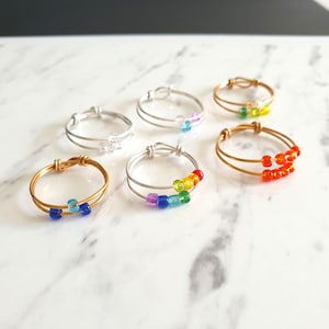 May include: A collection of six wire rings, each featuring a double-band design and small colourful beads. The rings are crafted from gold-tone and silver-tone wire, with beads in various shades including blue, green, yellow, purple, and orange.