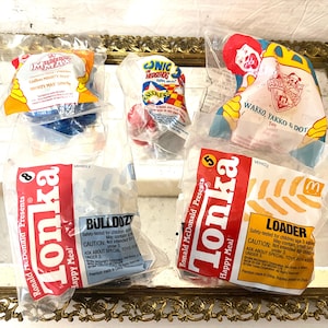 Peut inclure: Plusieurs jouets Happy Meal McDonald's scellés, dont des camions Tonka et des personnages de Sonic le Hérisson et de la série animée Wakko, Yakko & Dot. Les jouets sont dans leur emballage d'origine, avec des couleurs vives et des marques reconnaissables.