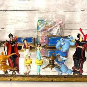 May include: A collection of Aladdin-themed figurines, including Aladdin, Jasmine, Genie, and Jafar. The figures are brightly colored and set on a mirrored surface, with a magic lamp and magic carpet. A sealed package is also visible.