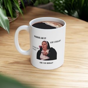 May include: White ceramic coffee mug with a handle, featuring a nun graphic and the phrases "WHERE AM I?", "AM I DEAD?", "IS THIS MY WAKE?", and "AM I IN HELL?". The mug is filled with dark coffee.