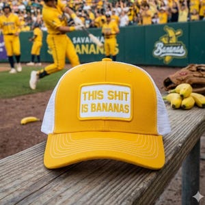 May include: Yellow and white baseball cap with the text "THIS SHIT IS BANANAS" on a patch. The cap is on a wooden bench with a baseball game in the background. A baseball glove and bananas are also visible.