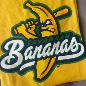 Puede incluir: Camiseta amarilla con el logo del equipo de b&eacute;isbol Savannah Bananas. El logo muestra un pl&aacute;tano con un bate de b&eacute;isbol y el nombre del equipo en letras blancas y verdes. La camiseta es de material suave.