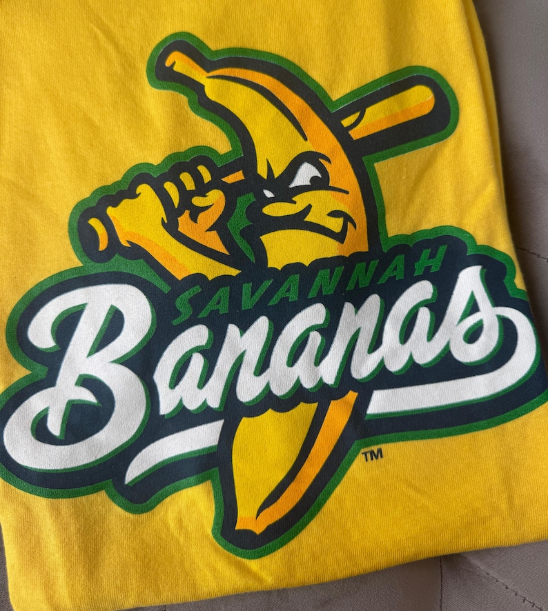 Puede incluir: Camiseta amarilla con el logo del equipo de b&eacute;isbol Savannah Bananas. El logo muestra un pl&aacute;tano con un bate de b&eacute;isbol y el nombre del equipo en letras blancas y verdes. La camiseta es de material suave.