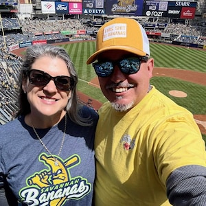 May include: A couple at a baseball game wearing sunglasses and apparel with the Savannah Bananas logo. One person wears a yellow hat with the text "THIS SHIT IS BANANAS". The stadium has large advertising signs.