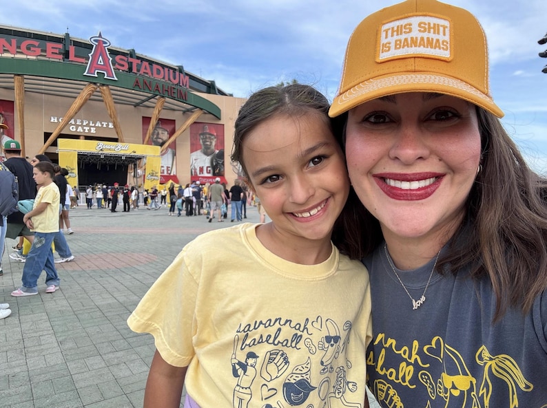 Puede incluir: Un selfie de dos personas sonriendo frente a un estadio. Una persona lleva una gorra de b&eacute;isbol amarilla con el texto "THIS SHIT IS BANANAS". La otra persona lleva una camiseta amarilla con el texto "savannah baseball."