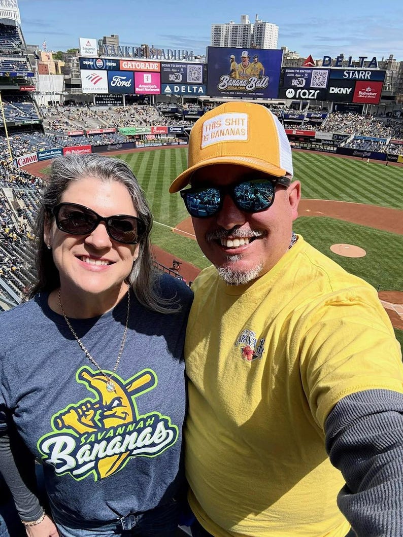 Puede incluir: Una foto de dos personas en un partido de b&eacute;isbol. Una persona lleva una camiseta amarilla y una gorra amarilla con el texto "THIS SHIT IS BANANAS". La otra persona lleva una camiseta azul con el logo de Savannah Bananas.