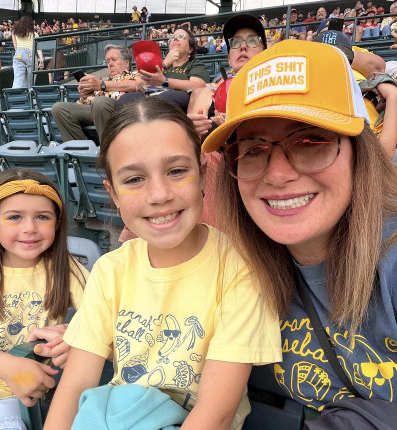 Puede incluir: Tres personas sonriendo en un partido de b&eacute;isbol. La mujer de enfrente lleva un sombrero amarillo con el texto "THIS SHIT IS BANANAS". Los dos ni&ntilde;os llevan camisetas amarillas con un dise&ntilde;o de b&eacute;isbol.