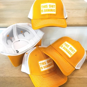 Puede incluir: Gorras de camionero amarillas y blancas con el texto "THIS SHIT IS BANANAS" en un parche rectangular. Las gorras tienen un panel frontal y una visera amarillos, con una parte trasera de malla blanca. Las gorras se muestran sobre una superficie de madera.