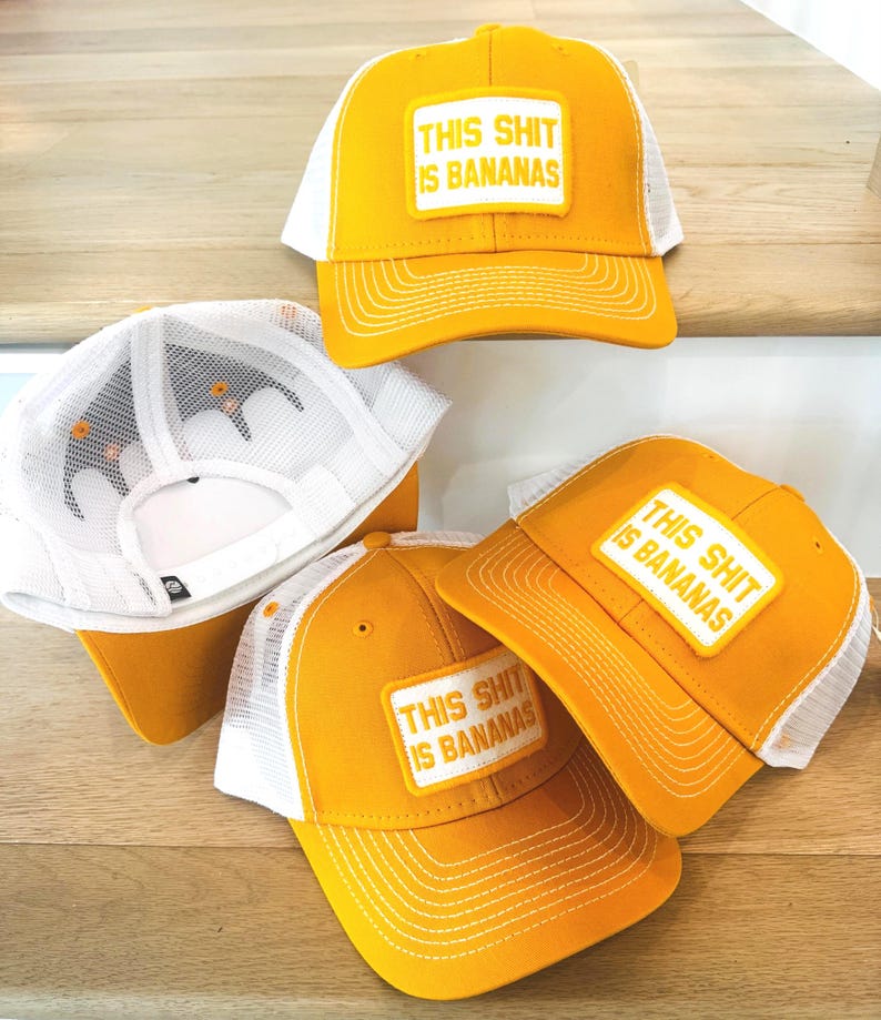 Puede incluir: Gorras de camionero amarillas y blancas con el texto "THIS SHIT IS BANANAS" en un parche rectangular. Las gorras tienen un panel frontal y una visera amarillos, con una parte trasera de malla blanca. Las gorras se muestran sobre una superficie de madera.