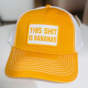 Puede incluir: Gorra de camionero amarilla con la parte trasera de malla blanca y visera curva. La gorra presenta un parche rectangular blanco con la frase "THIS SHIT IS BANANAS" en letras may&uacute;sculas.