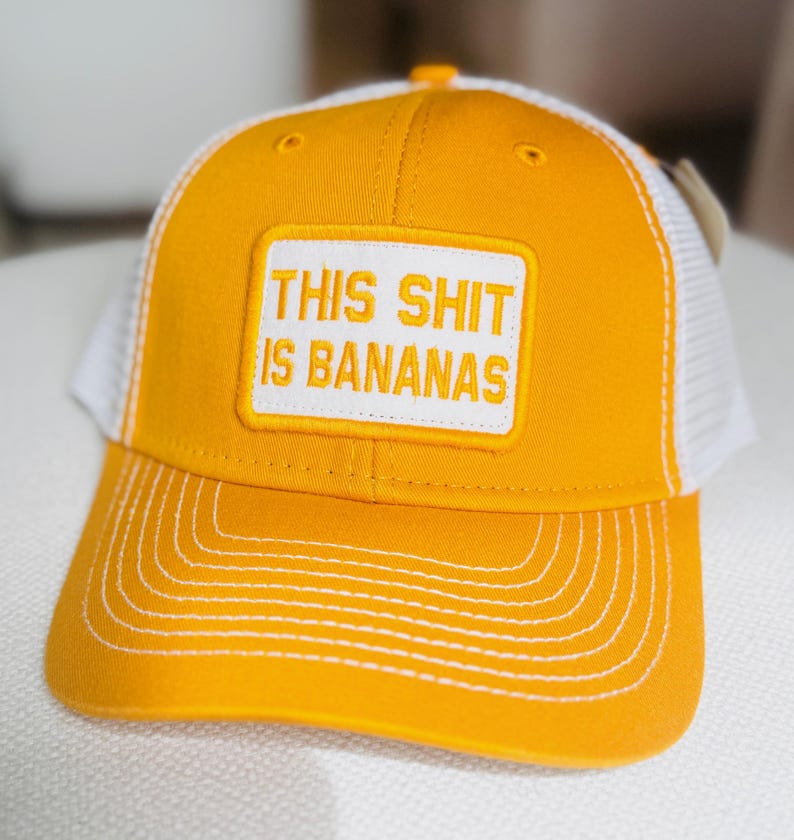 Puede incluir: Gorra de camionero amarilla con la parte trasera de malla blanca y visera curva. La gorra presenta un parche rectangular blanco con la frase "THIS SHIT IS BANANAS" en letras may&uacute;sculas.