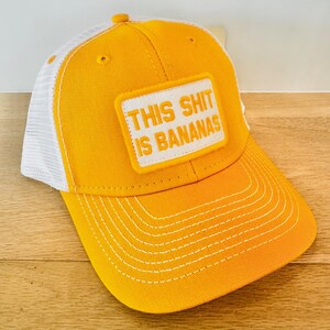 Puede incluir: Gorra de b&eacute;isbol amarilla con la parte trasera de malla blanca. La gorra presenta el texto "THIS SHIT IS BANANAS" en un parche rectangular. Las costuras blancas acent&uacute;an la visera y la parte delantera de la gorra.